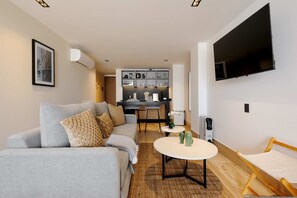 Una sala de estar moderna con un sofá, una pequeña mesa redonda, un televisor montado en la pared y una zona de cocina al fondo.