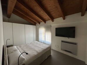 Ein Schlafzimmer mit einem Bett, einem an der Wand montierten Fernseher und einem Fenster mit Vorhängen.