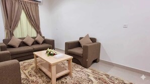 A living room with a sofa, armchair, and coffee table, a patterned rug, and a window with curtains. - Delta Park Serviced Apartments (Riyadh)