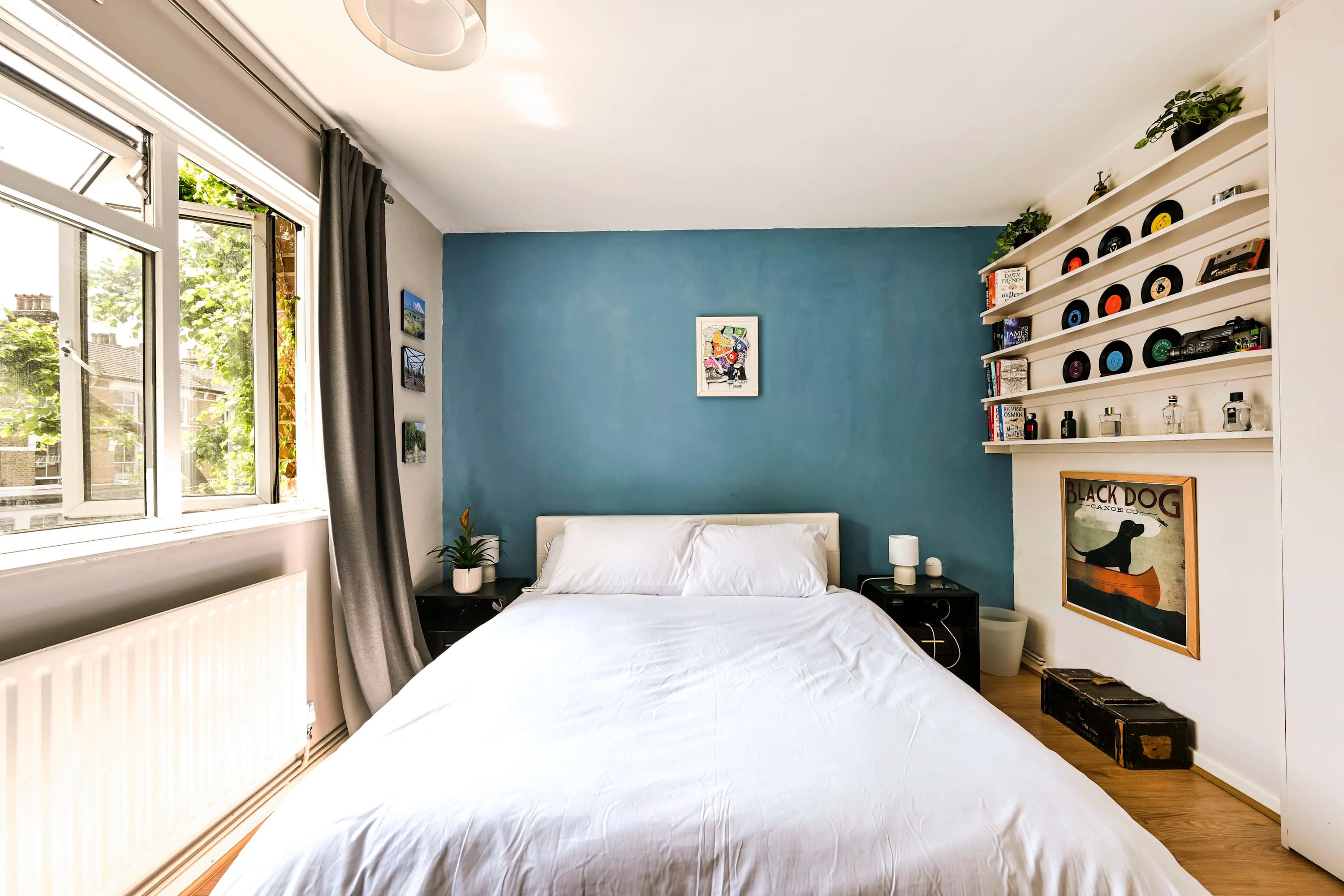 Un dormitorio con cama, ventana, estante lleno de libros y discos, y un cuadro enmarcado.