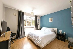 Ein Schlafzimmer mit Bett, Schreibtisch mit Computer, Stuhl, Nachttisch mit Lampe und einem Fenster mit Vorhängen.