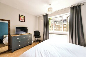 Ein Schlafzimmer mit Bett, Kommode, Stuhl, Fernseher und einem Fenster mit Vorhängen.