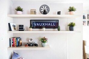 Eine Wand mit drei Regalen, auf denen unterschiedliche Gegenstände stehen. Das oberste Regal zeigt Blumentöpfe, eine Uhr und ein gerahmtes Bild. Im mittleren Regal befindet sich ein großes Schild mit 'VAUXHALL' sowie ein gerahmtes Foto. Das unterste Regal enthält Bücher, eine Kamera und eine Vase mit Blumen.