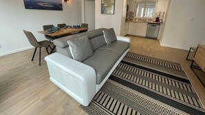En moderne stue med en grå sofa, spisebord og stole, samt et køkken i baggrunden og gulvtæpper med mønster.