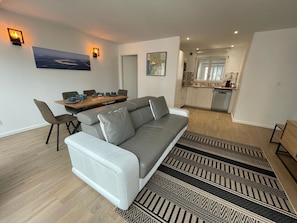 Ein modernes Wohnzimmer mit grauem Sofa, Esstisch und Stühlen, sowie einer Küche im Hintergrund und gemusterten Teppichen.