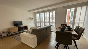 En moderne stue med sofa, spisebord med stole, fjernsyn og store vinduer.