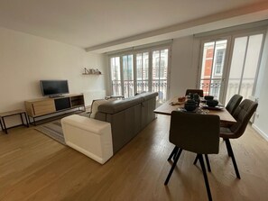 Ein modernes Wohnzimmer mit Sofa, Esstisch mit Stühlen, Fernseher und großen Fenstern.