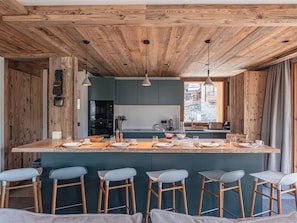 A kitchen with wooden ceiling beams, a central island with bar stools, and a view of a stone building outside. - Ski-in/ski-out apartment, sunny view - 11 guests (Les Allues)