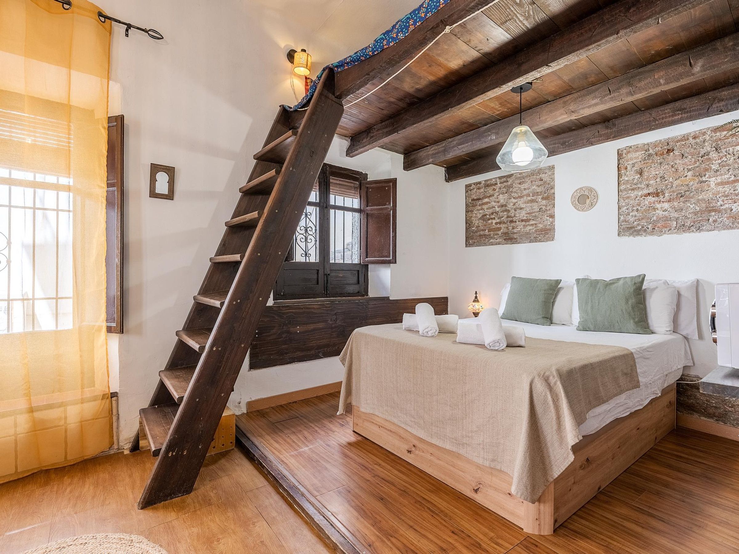 A bedroom with a wooden bed, a ladder, a window with curtains, and a brick wall.