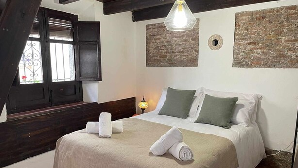 A bedroom with a bed, pillows, a nightstand, a lamp, a window with shutters, and a brick wall. - Bright studio in Albaicín - 2 guests (Granada)