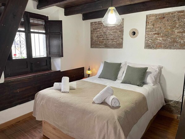 Ein Schlafzimmer mit einem Bett, Kissen, einem Nachttisch, einer Lampe, einem Fenster mit Fensterläden und einer Ziegelwand.