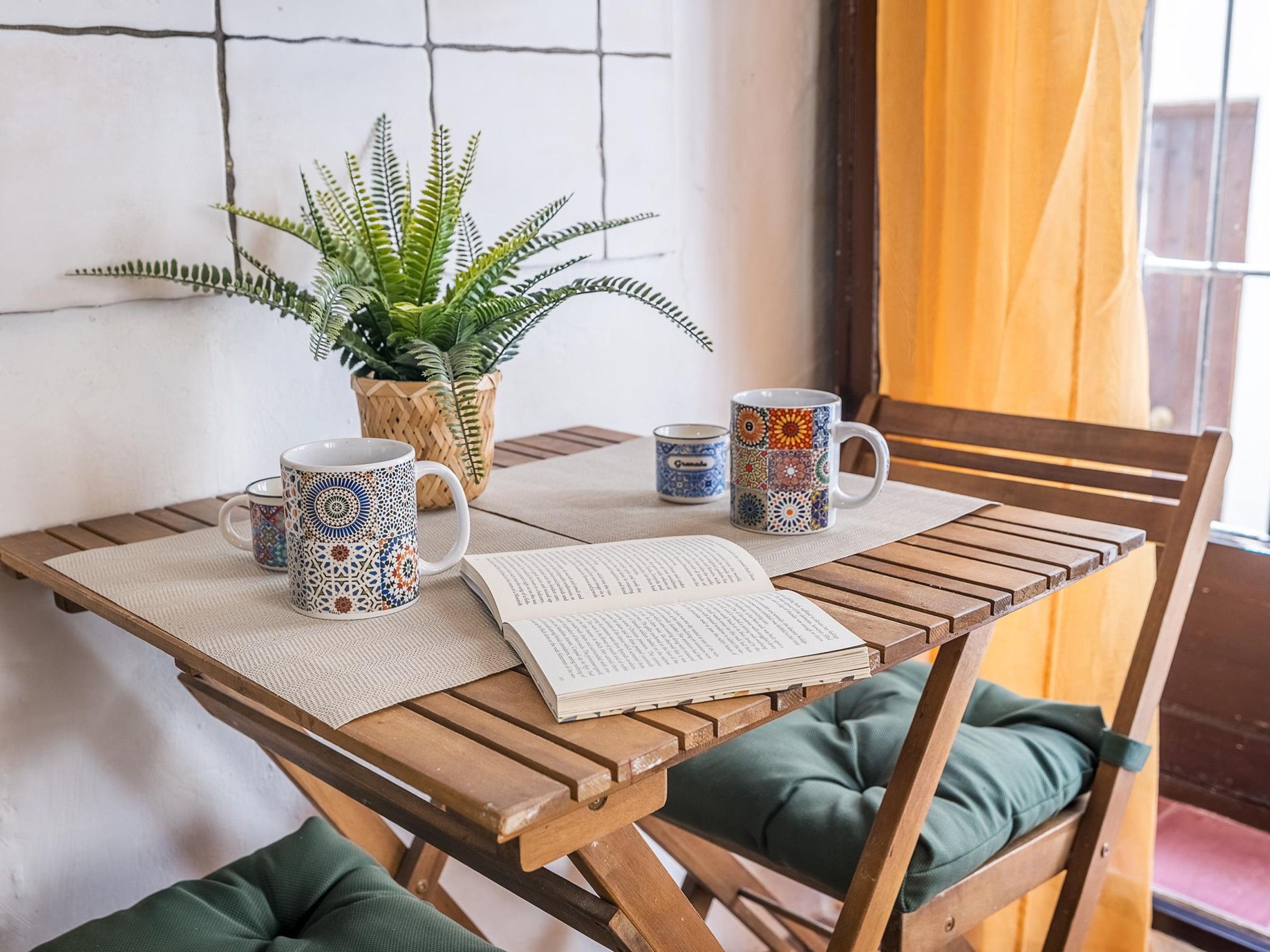 Een houten tafel met een geweven placemat, twee decoratieve mokken, een pot met varen en een open boek.