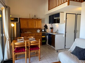 A compact kitchen with wooden cabinets, a dining table with chairs, a bed with pillows, and a small refrigerator. - Apartment with sea view, pool, parking - 6 guests (Gruissan)