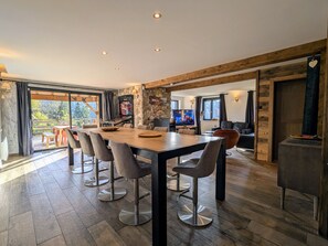 A spacious dining area with a long table and modern chairs, a stone accent wall, and a television in the background. - House panoramic view, parking - 2 guests (Gérardmer)