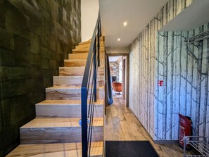Eine moderne Treppe mit Holzhäuschen und Metallgeländer, die zu einem Raum mit Steinwand und einem an der Wand befestigten Feuerlöscher führt.