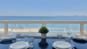 Mesa de comedor con platos, vasos y cubiertos, con vista a un mar azul y terreno lejano.