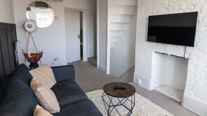 Een woonkamer met een bank, een ronde salontafel, een aan een bakstenen muur bevestigde televisie en een gang die naar een andere kamer leidt.