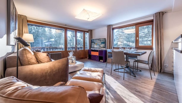 Una sala de estar moderna con un sofá, una mesa de comedor con sillas, un televisor y ventanas amplias con vistas a árboles.