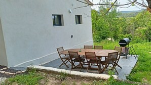 Ein Essbereich im Freien mit einem runden Tisch und Stühlen, einem Grill und einem weißen Gebäude mit Fenstern.