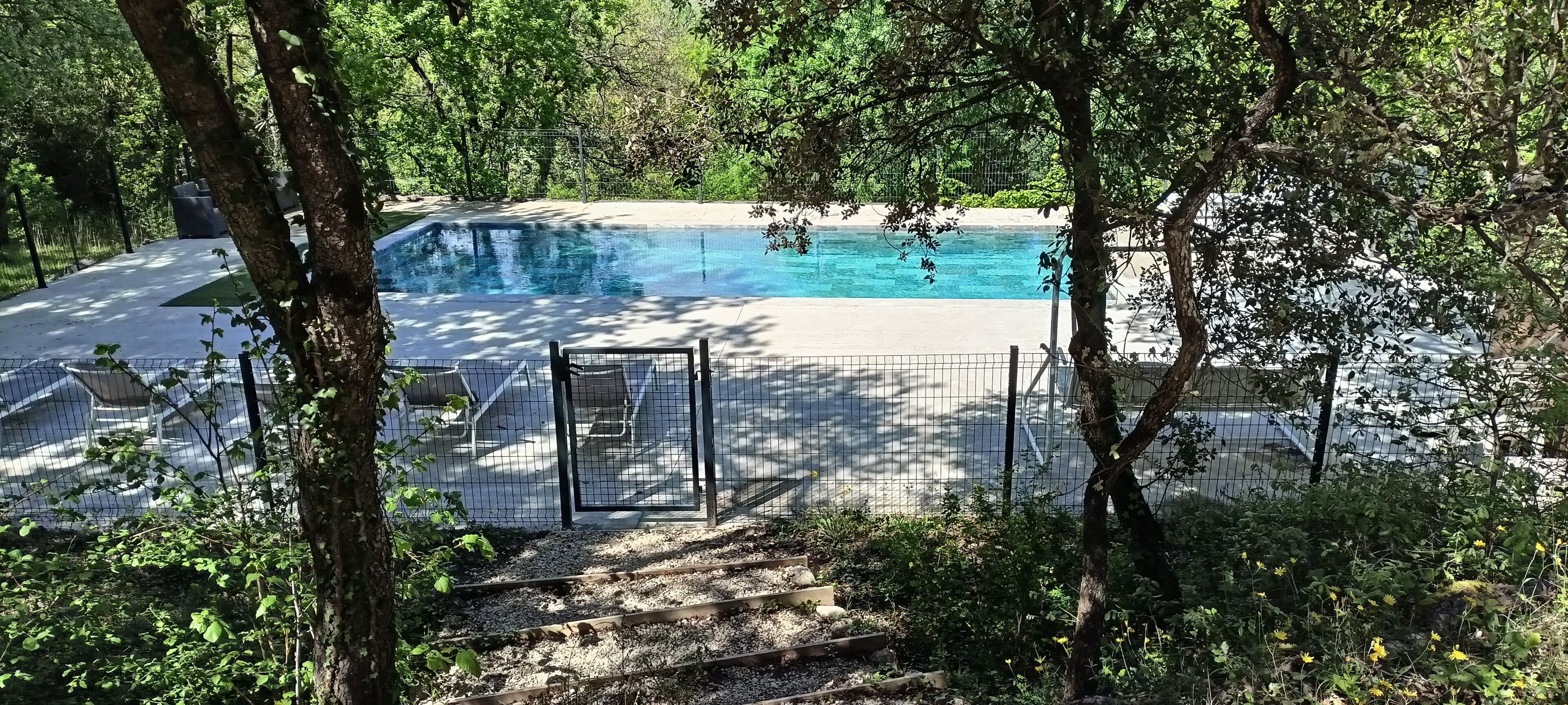 Une piscine entourée d’arbres, avec une clôture et des marches menant à l’espace piscine.