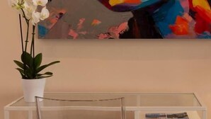 A painting of a woman with a colorful abstract background, a white orchid in a white pot, and a clear glass table with a magazine. - Room Boutique 32 (Dénia)