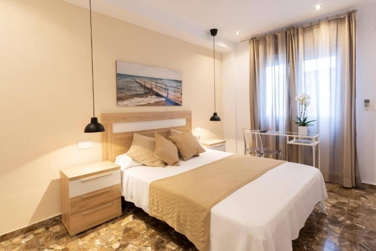 Habitación de hotel con cama, mesitas de noche, escritorio, silla, un cuadro en la pared y ventana con cortinas.