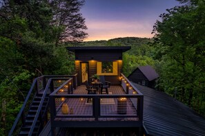Eine Holzterrasse mit Tisch und Stühlen, umgeben von Bäumen, und ein kleines Gebäude mit dunklem Dach.
