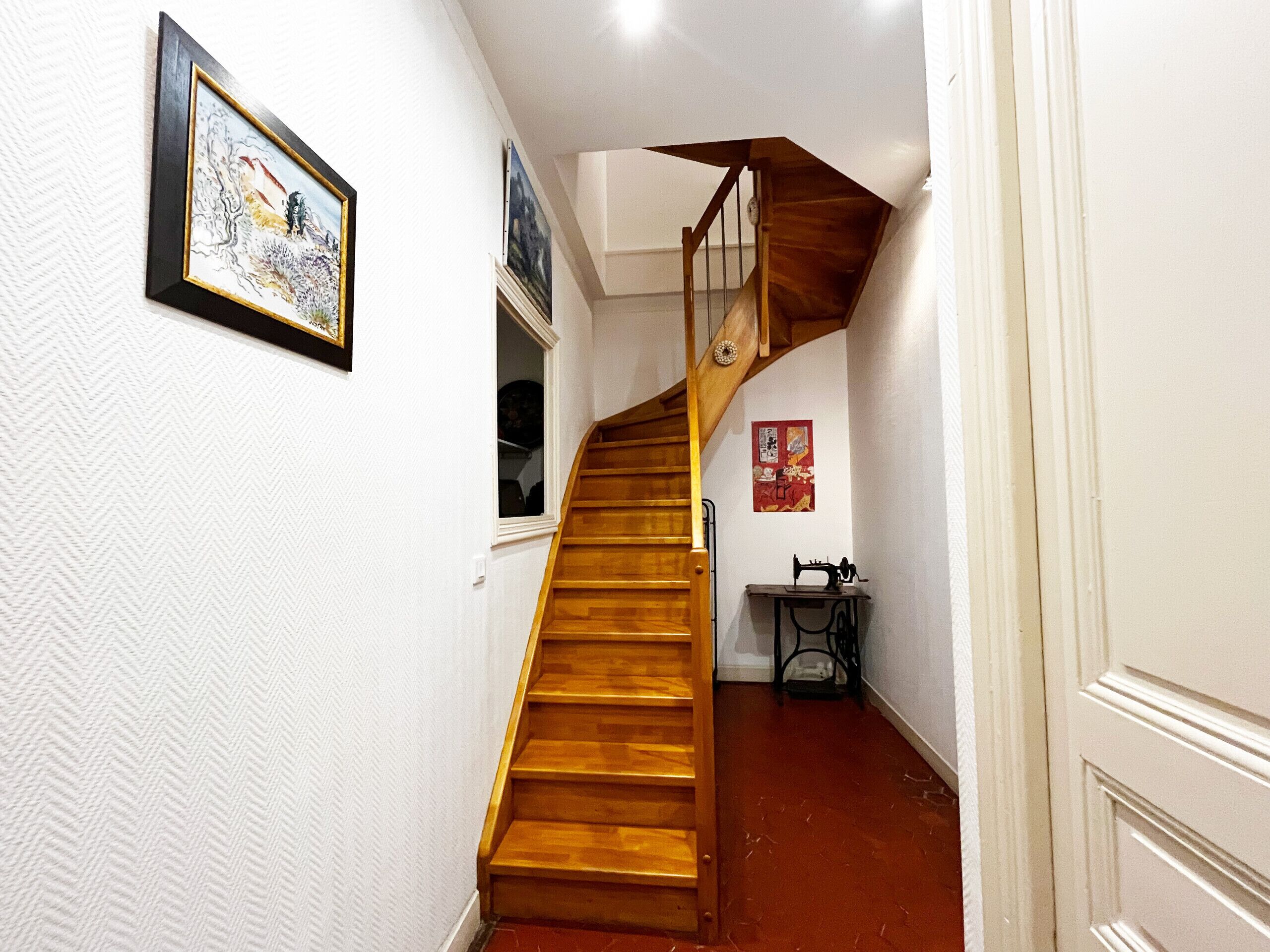 Un escalier en bois menant à un deuxième étage, des murs blancs et une petite table sur laquelle se trouve une machine à coudre.