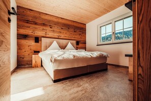 A bedroom with a wooden ceiling, a bed with a headboard, a window with a view of trees, and a bedside table.