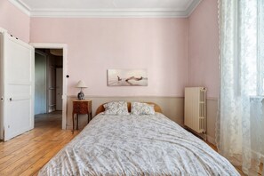 A bedroom with a bed, a bedside table, a lamp, a radiator, and a door leading to another room.