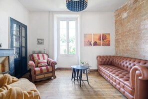 A room with a wooden floor, a brown leather sofa, a striped armchair, a small round table, a blue lamp, and a brick wall.