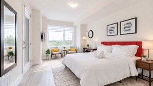 Ein Hotelzimmer mit einem großen Bett, zwei Stühlen, einem kleinen Tisch, einem roten Kopfteil und Blick durch das Fenster.