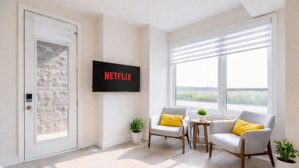 Ein modernes Wohnzimmer mit einem Flachbildfernseher, zwei Sesseln, einem kleinen Tisch und Blick durch große Fenster.