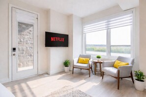 Un moderno salón con televisor de pantalla plana, dos sillones, una mesita y vistas amplias a través de grandes ventanales.