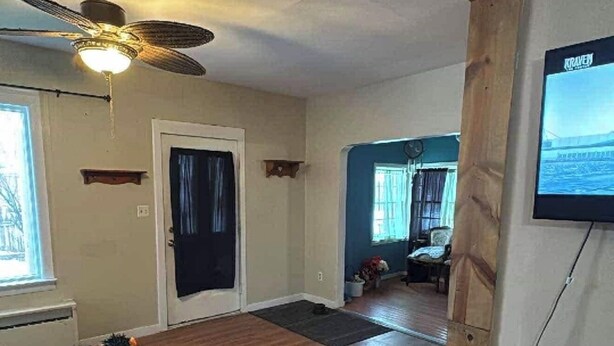 Een ruime woonkamer met houten vloeren, een plafondventilator, een deur en een aan de muur bevestigde televisie.
