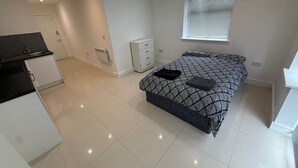 A modern bedroom with a bed, a small bedside table, a window with blinds, and a kitchenette area with cabinets and a countertop. - Radstock Road - Room 2 (Southampton)