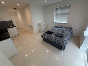 A modern bedroom with a bed, a small bedside table, a window with blinds, and a kitchenette area with cabinets and a countertop.