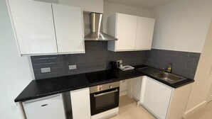 A compact kitchen with white cabinets, a black countertop, a stainless steel range hood, a built-in oven, and a sink. - Radstock Road - Room 2 (Southampton)