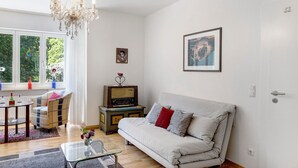 Een woonkamer met een bank, een televisie, een kroonluchter en een raam met uitzicht op bomen.