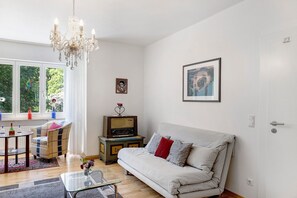 Ein Wohnzimmer mit einer Couch, einem Fernseher, einer Chandelüster und einem Fenster mit Blick auf Bäume.