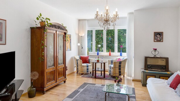Een woonkamer met een televisie, een houten kast, een kroonluchter, een bank, een tafel en een raam met uitzicht op groen.