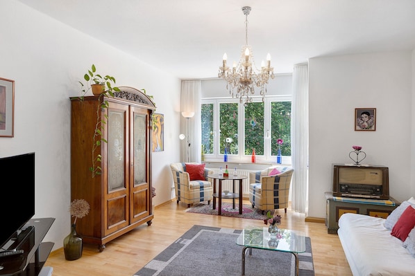 A living room with a TV, a wooden cabinet, a chandelier, a sofa, a table, and a window with a view of greenery.