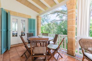 Une terrasse couverte avec une table et des chaises en bois, une porte blanche avec des volets bleus et une vue sur la verdure.