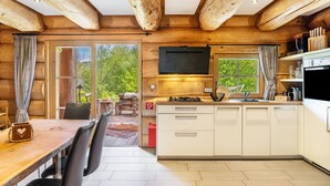 A log cabin interior with a wooden dining table, chairs, a kitchen with white cabinets, a TV mounted above the stove, and a view of greenery outside. - Chalet "Weiss" with Mountain Views, Private Garden, and Wi-Fi (Wörgl)
