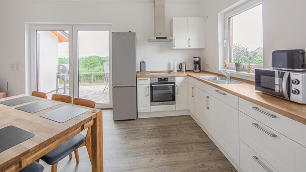 A modern kitchen with a refrigerator, oven, microwave, and sink. There is a dining table with chairs and a window with a view. - Ferienwohnungen in Filz in der Eifel (Filz)