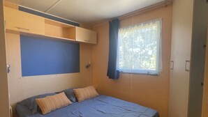 A small, single-bed room with a built-in headboard, a window with curtains, and a ceiling light.