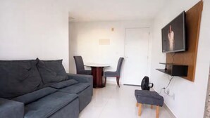 Una sala de estar con un sofá, una mesa de comedor con sillas, un televisor montado en la pared y un pequeño banco.