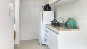 Una cocina compacta con gabinetes blancos, un refrigerador blanco, una encimera con cafetera, hervidor y un cubo de basura verde.