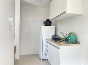 A compact kitchen with white cabinets, a white refrigerator, a countertop with a coffee maker, a kettle, and a green trash bin. - Fully furnished apartment 5 min from Recreio Beach, Rio de Janeiro (Rio de Janeiro)