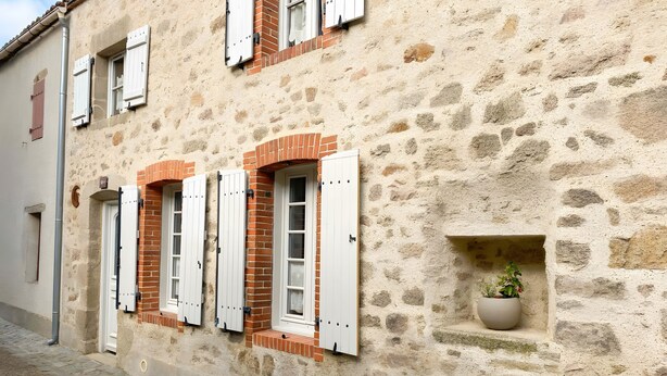 Un edificio de piedra con contraventanas blancas, una maceta pequeña con plantas y una calle adoquinada.